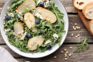 Photo of Tasty salad with arugula, pear, blue cheese, peanuts and blueberries in bowl on wooden table, flat lay