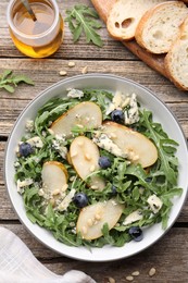 Photo of Tasty salad with arugula, pear, blue cheese, peanuts and blueberries in bowl on wooden table, flat lay