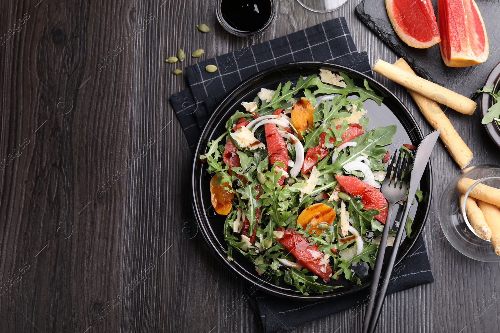 Photo of Tasty salad with arugula, cheese, grapefruit, vegetable and balsamic sauce served on dark wooden table, flat lay. Space for text