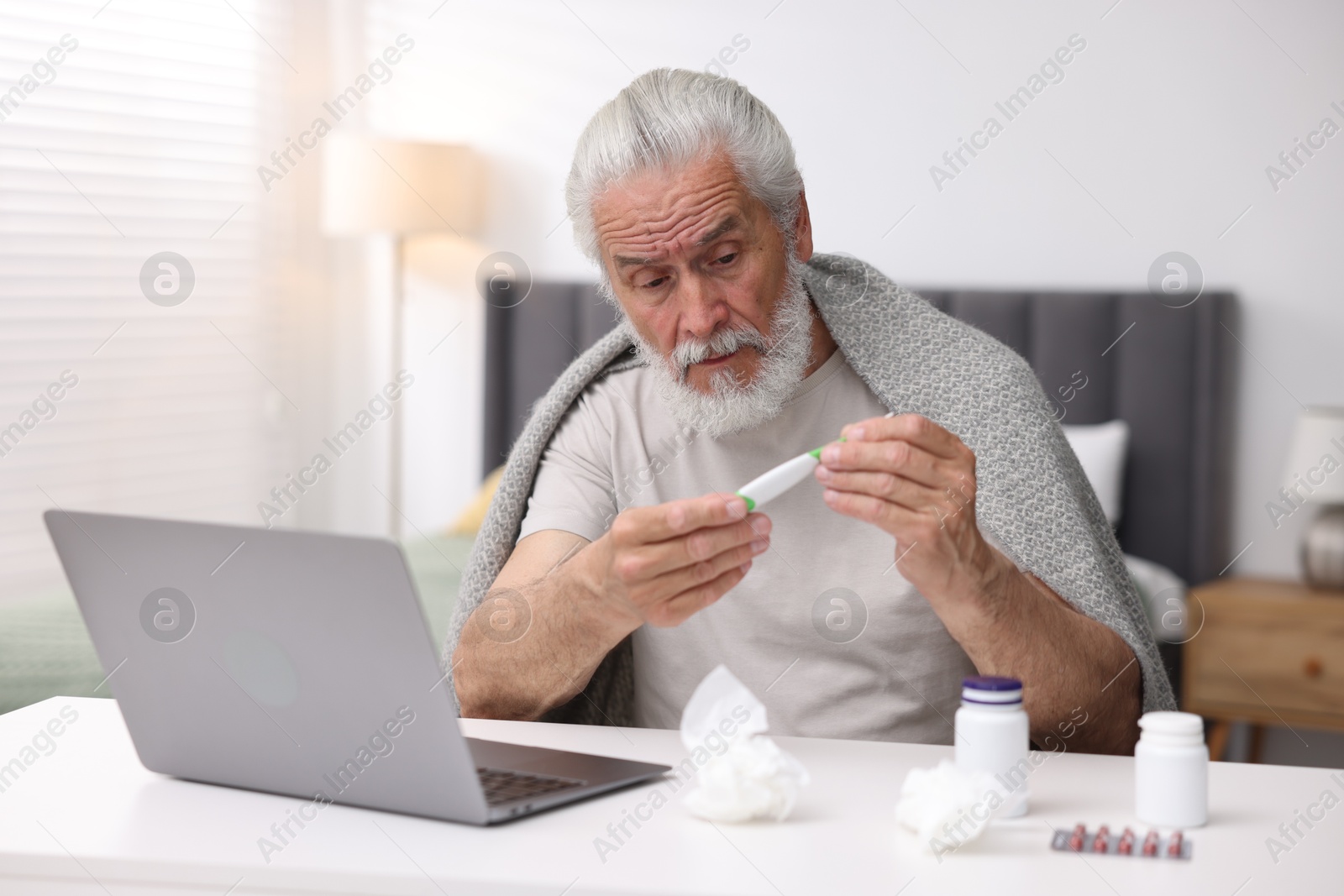 Senior man with thermometer having online consultation with doctor via laptop at table indoors Photo of Senior man with thermometer having online consultation with doctor via laptop at table indoors