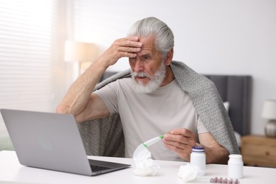 Photo of Senior man with thermometer having online consultation with doctor via laptop at table indoors