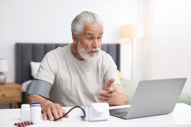 Photo of Senior man measuring blood pressure while having online consultation with doctor via laptop at table indoors
