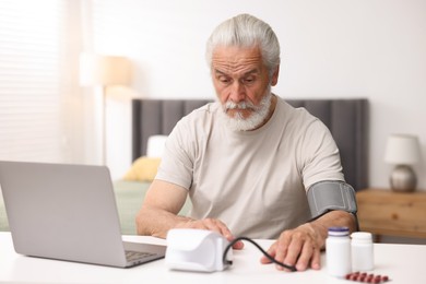 Photo of Senior man measuring blood pressure while having online consultation with doctor via laptop at table indoors