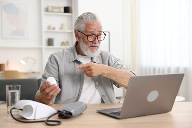 Photo of Senior man with pills having online consultation with doctor via laptop at table indoors