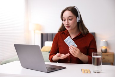 Photo of Woman in headphones with pills having online consultation with doctor via laptop at table indoors