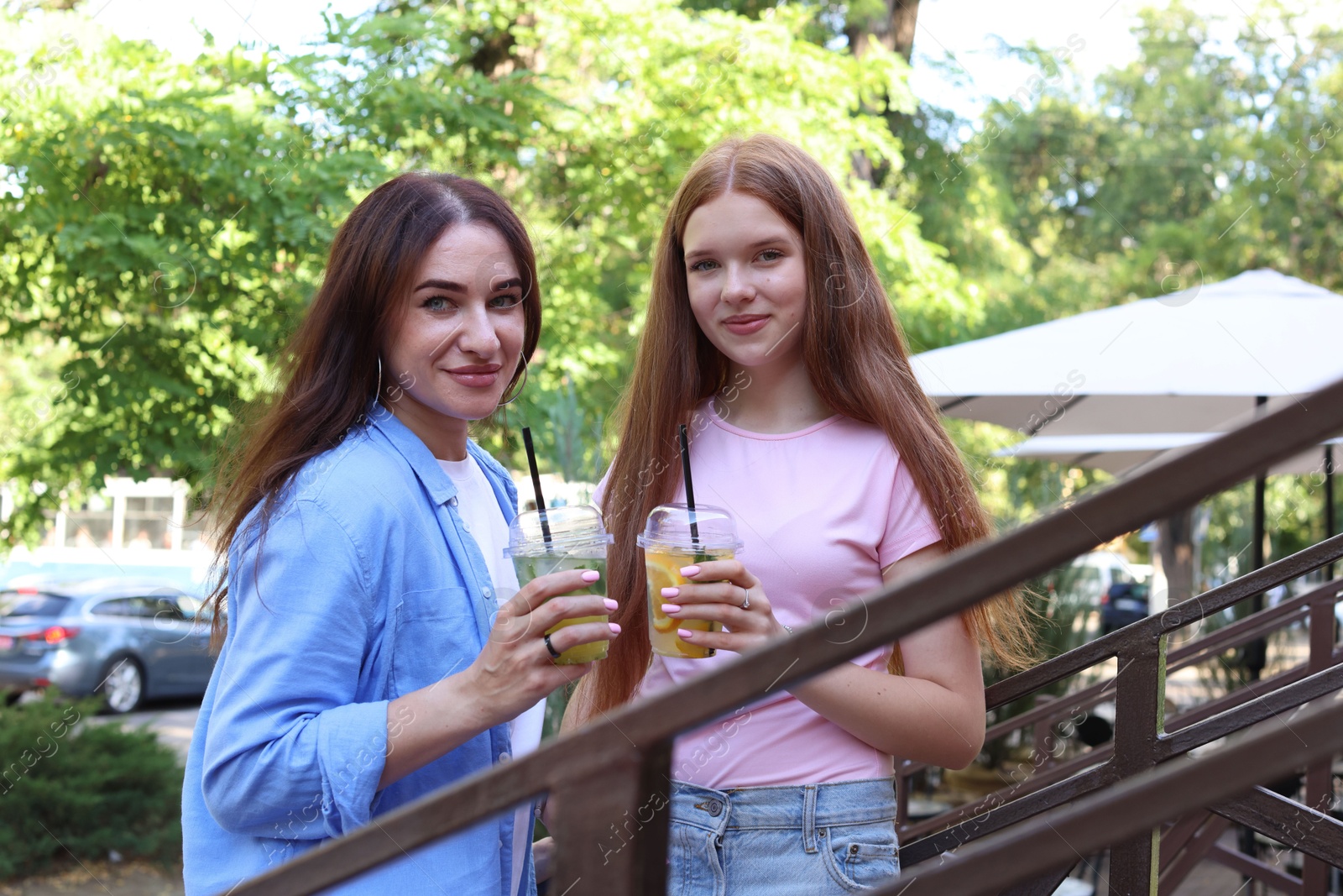 Photo of Portrait of beautiful mother and her teenage daughter with refreshing drinks on stairs outdoors