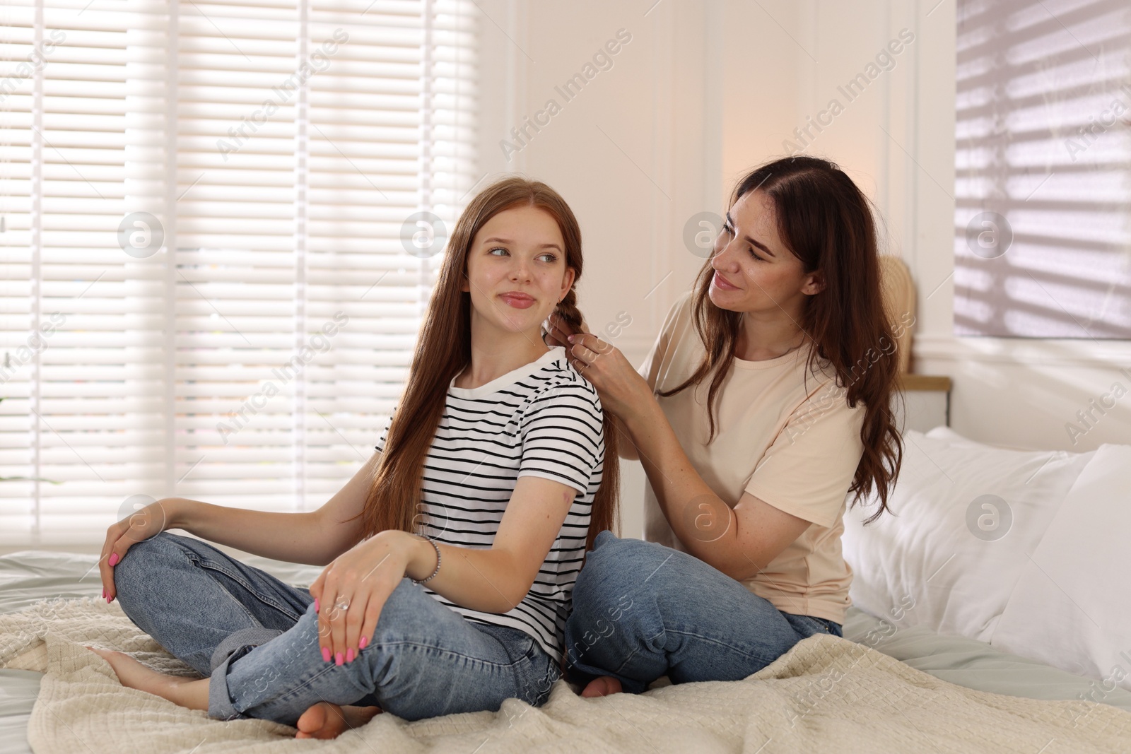 Photo of Mother braiding her teenage daughter's hair on bed at home