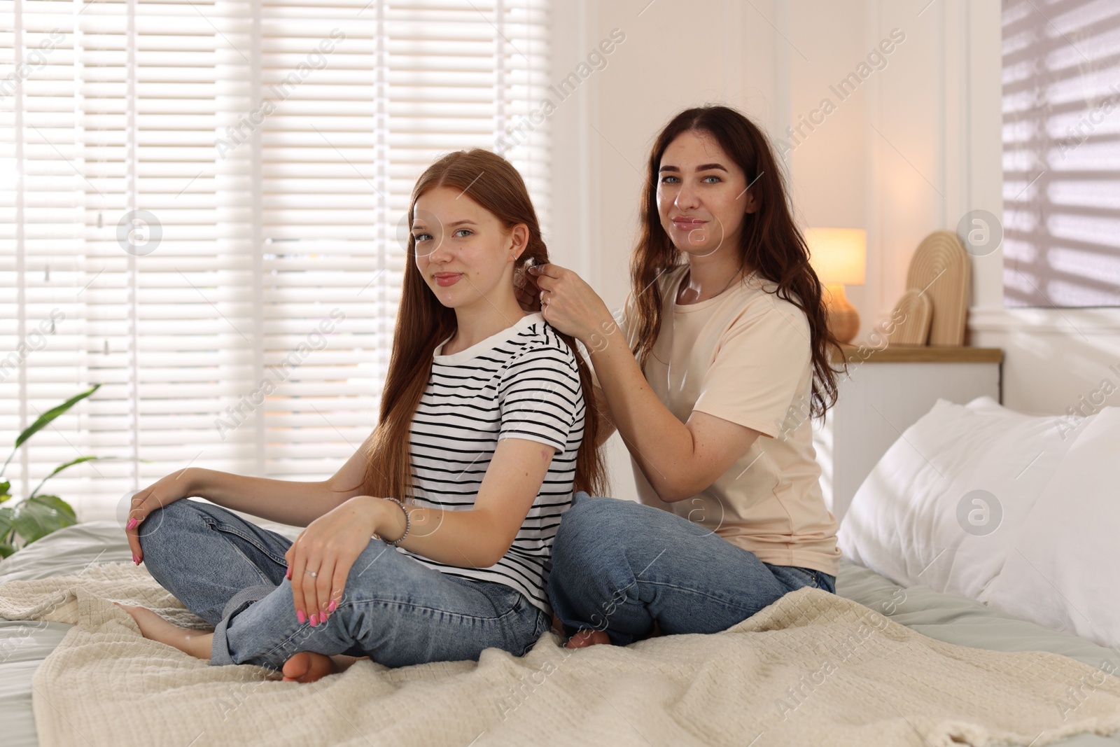 Photo of Mother braiding her teenage daughter's hair on bed at home