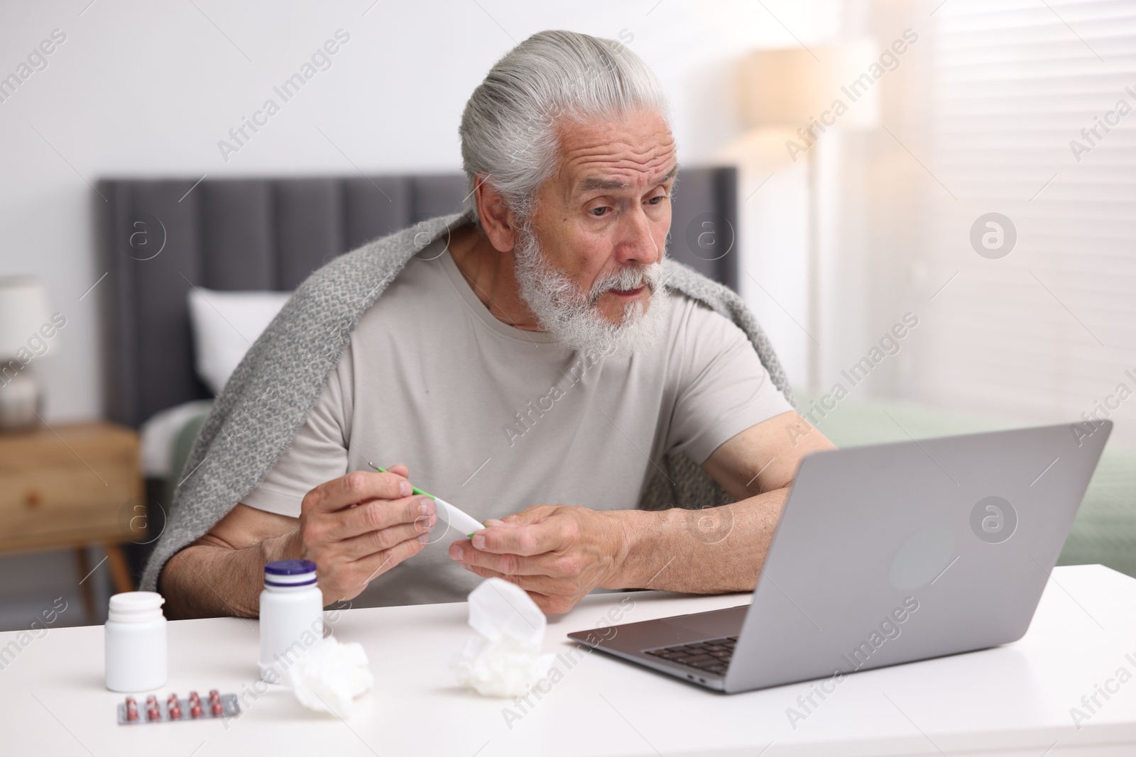 Senior man with thermometer having online consultation with doctor via laptop at table indoors Photo of Senior man with thermometer having online consultation with doctor via laptop at table indoors