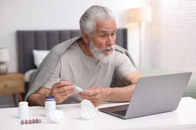 Photo of Senior man with thermometer having online consultation with doctor via laptop at table indoors