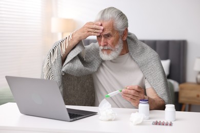 Photo of Senior man with thermometer having online consultation with doctor via laptop at table indoors