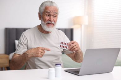 Photo of Senior man with pills having online consultation with doctor via laptop at table indoors