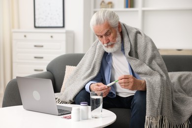 Photo of Senior man with thermometer having online consultation with doctor via laptop at home