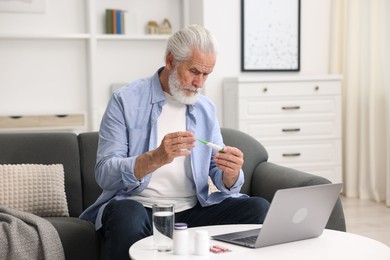 Photo of Senior man with thermometer having online consultation with doctor via laptop at home