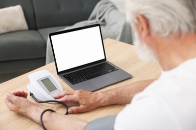 Photo of Senior man measuring blood pressure while having online consultation with doctor via laptop at table indoors, closeup