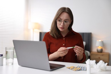 Photo of Woman with thermometer having online consultation with doctor via laptop at table indoors