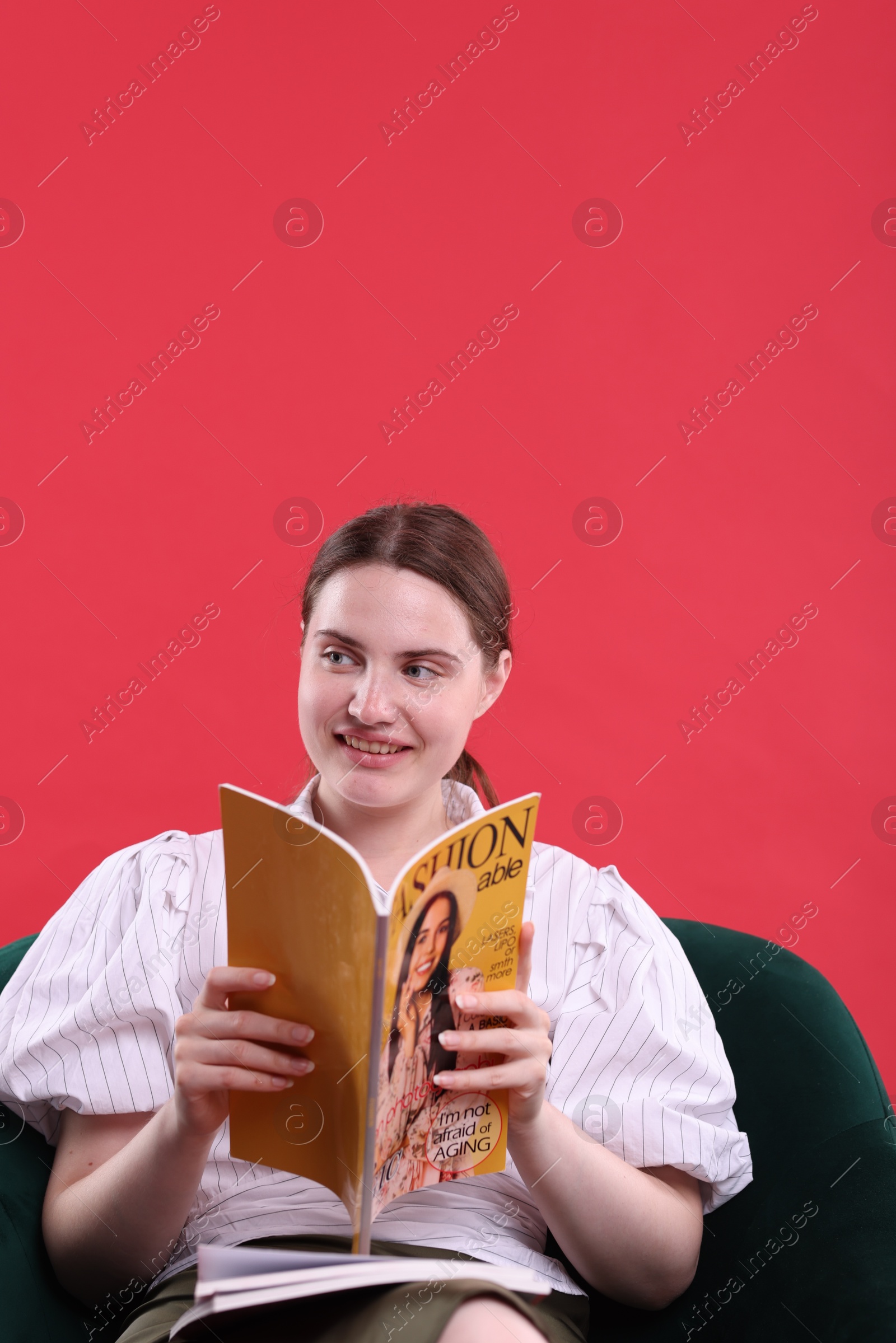 Woman with fashion magazine on armchair against red background, space for text Photo of Woman with fashion magazine on armchair against red background, space for text