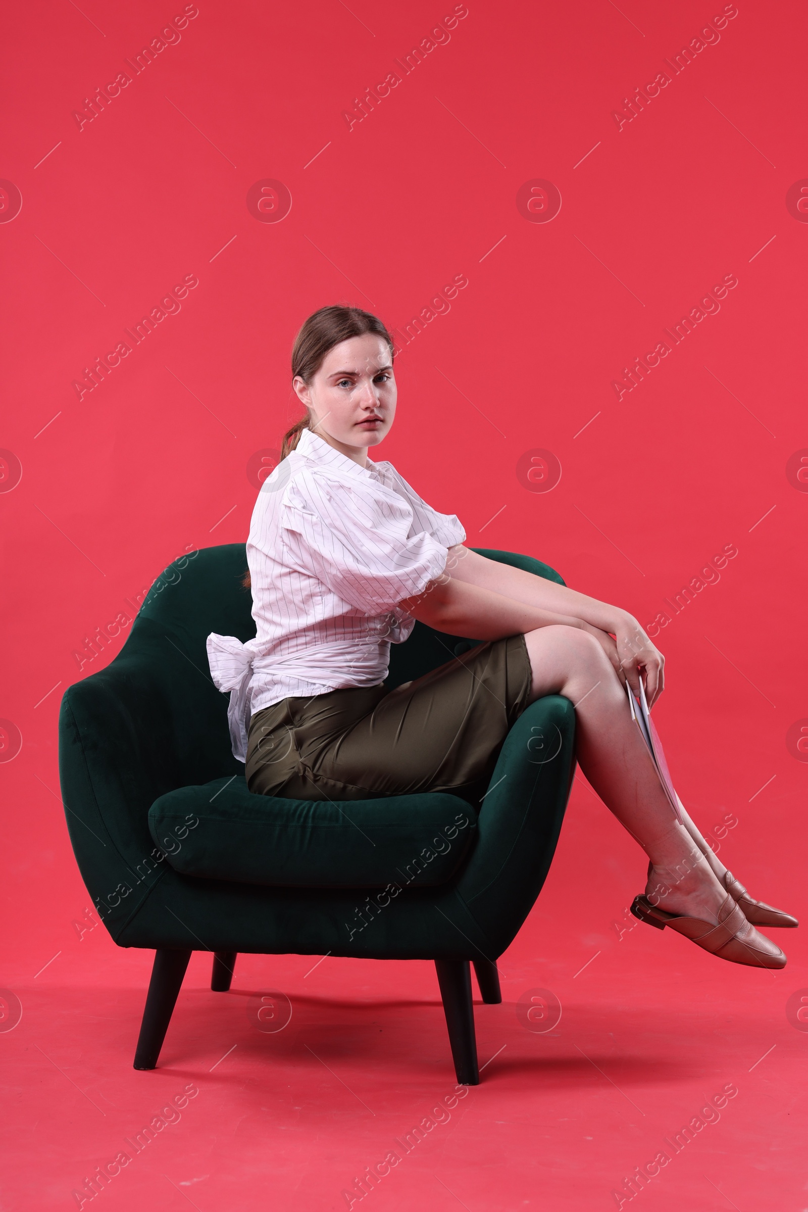 Beautiful young woman sitting on armchair against red background Photo of Beautiful young woman sitting on armchair against red background