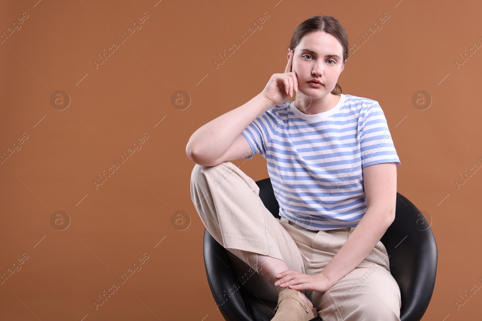 Beautiful young woman sitting on armchair against brown background, space for text Photo of Beautiful young woman sitting on armchair against brown background, space for text