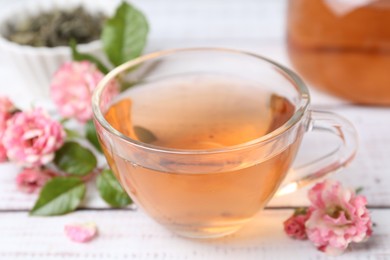 Photo of Tasty rose tea in glass cup and flowers on white wooden table, closeup