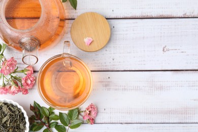 Photo of Tasty rose tea in glass cup, teapot, flowers and leaves on light wooden table, flat lay. Space for text