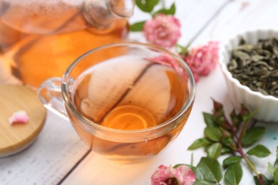 Photo of Tasty rose tea in glass cup, flowers and leaves on white wooden table, closeup