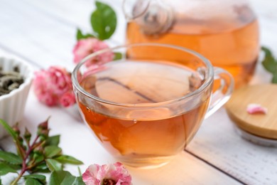 Photo of Tasty rose tea in glass cup, flowers and leaves on white wooden table, closeup