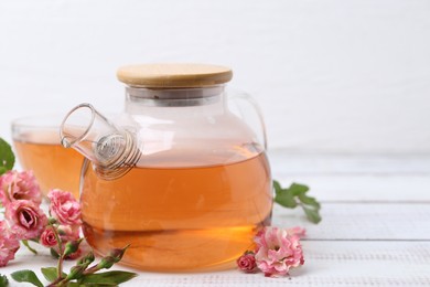 Photo of Tasty rose tea in glass teapot, flowers and leaves on white wooden table, closeup