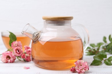 Photo of Tasty rose tea in glass teapot, flowers and leaves on white wooden table, closeup