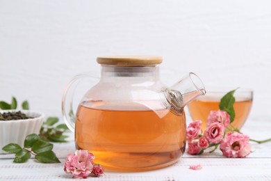 Photo of Tasty rose tea in glass teapot, flowers and leaves on white wooden table, closeup