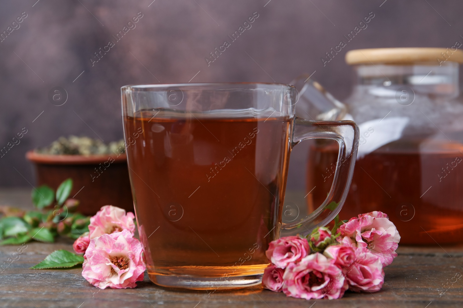 Tasty rose tea in glass cup and flowers on wooden table against brown background, closeup Photo of Tasty rose tea in glass cup and flowers on wooden table against brown background, closeup