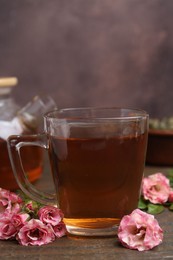 Photo of Tasty rose tea in glass cup and flowers on wooden table against brown background, closeup