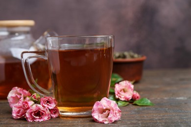 Photo of Tasty rose tea in glass cup and flowers on wooden table against brown background, closeup