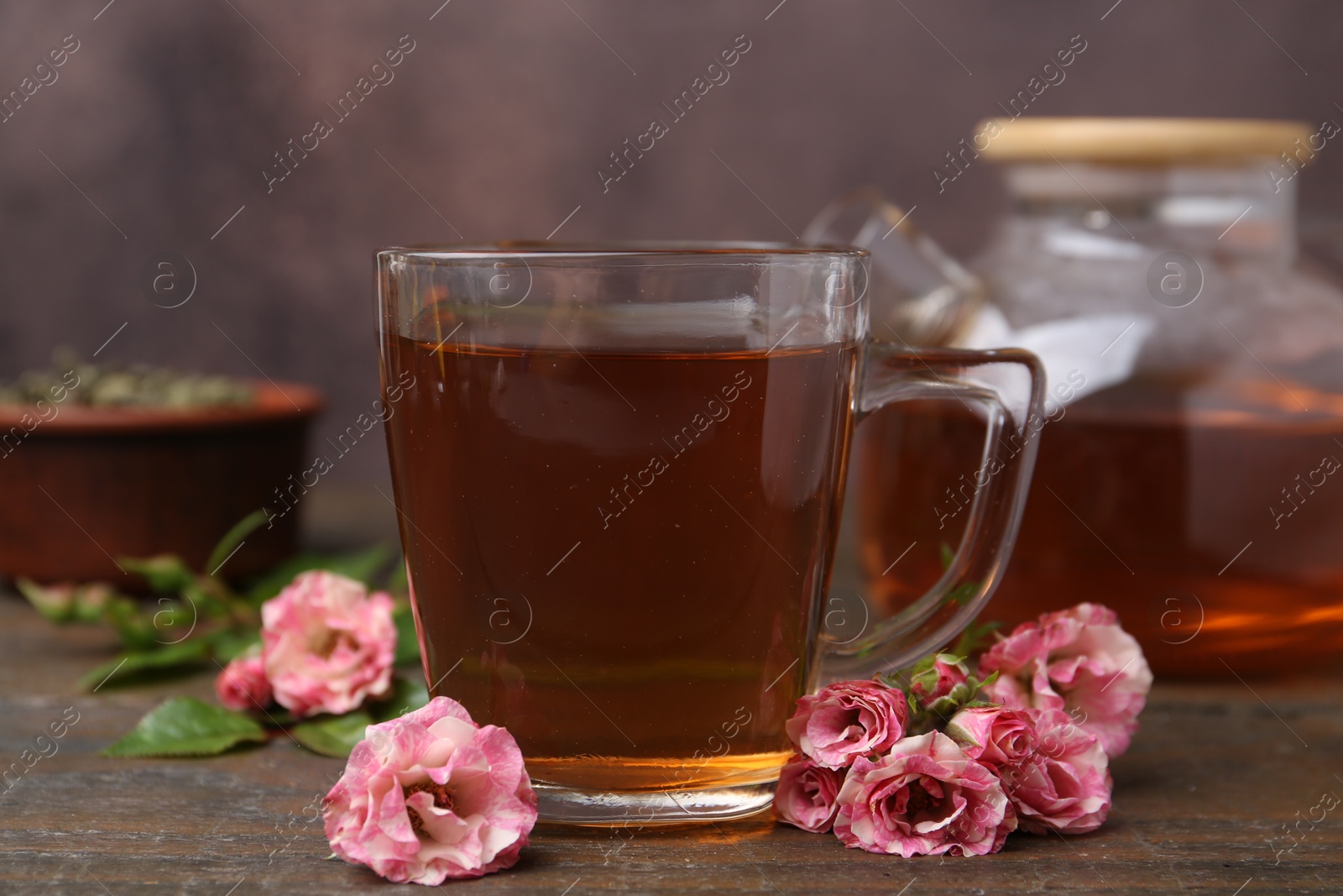 Tasty rose tea in glass cup and flowers on wooden table against brown background, closeup Photo of Tasty rose tea in glass cup and flowers on wooden table against brown background, closeup