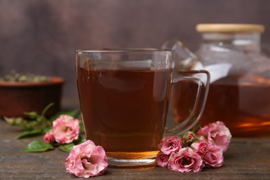 Photo of Tasty rose tea in glass cup and flowers on wooden table against brown background, closeup