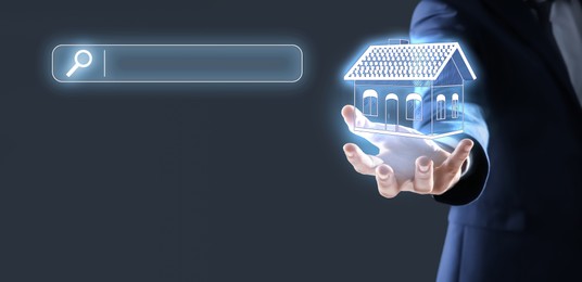 Image of Real estate holding virtual house against dark grey background with search bar, closeup. Searching for housing online