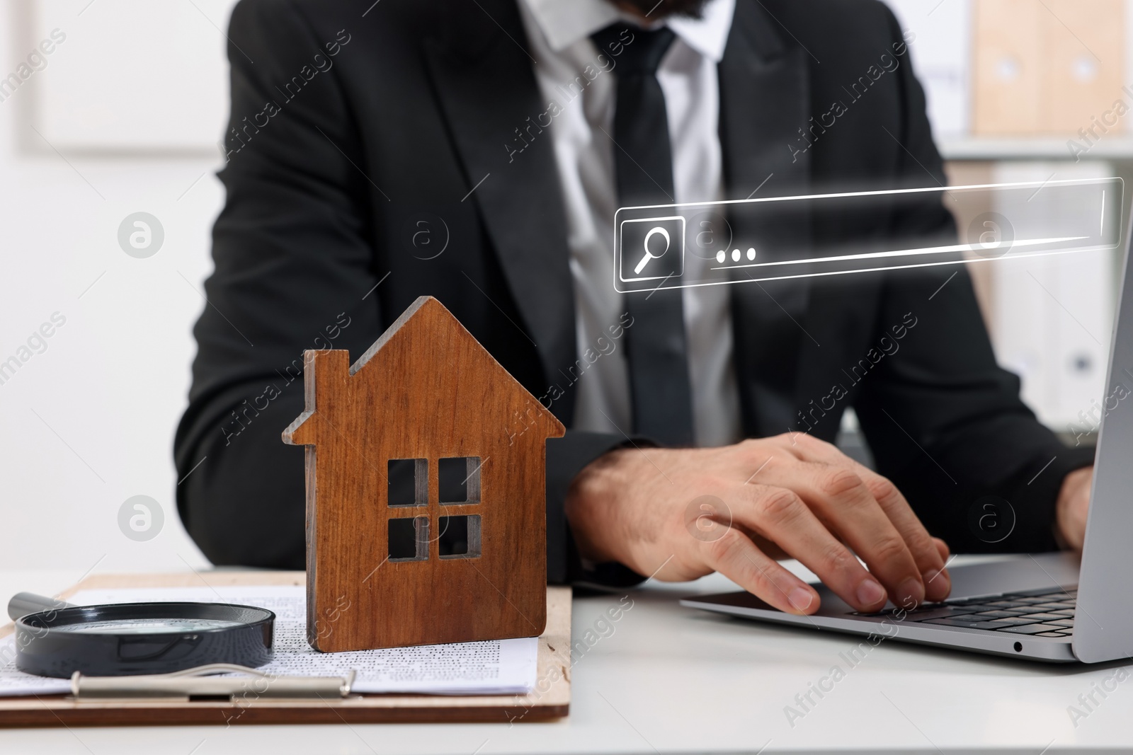 Searching for housing online. Real estate agent using laptop at table with magnifying glass and house model, closeup. Search bar above computer Image of Searching for housing online. Real estate agent using laptop at table with magnifying glass and house model, closeup. Search bar above computer