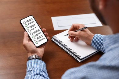 Image of Man passing online test on his mobile phone and writing in notebook at table, closeup