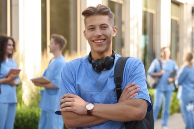 Photo of Happy medical student in uniform with backpack and headphones near building outdoors, selective focus