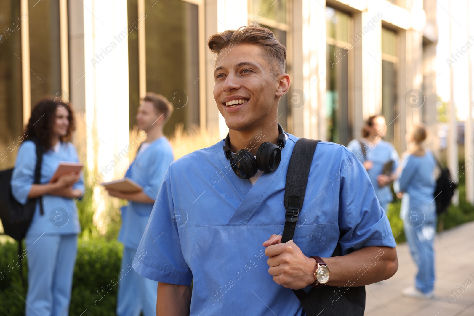 Photo of Happy medical student in uniform with backpack and headphones near building outdoors, selective focus