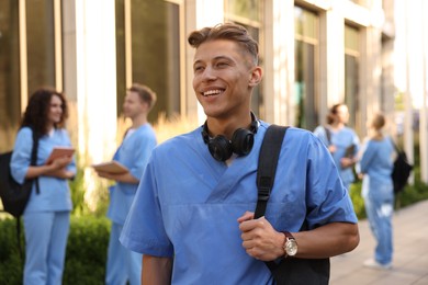 Photo of Happy medical student in uniform with backpack and headphones near building outdoors, selective focus