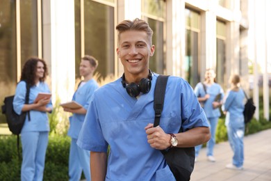 Photo of Happy medical student in uniform with backpack and headphones near building outdoors, selective focus