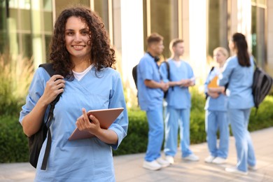 Photo of Happy medical student in uniform with backpack and tablet near building outdoors, selective focus