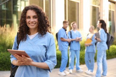 Photo of Happy medical student in uniform with backpack and tablet near building outdoors, selective focus