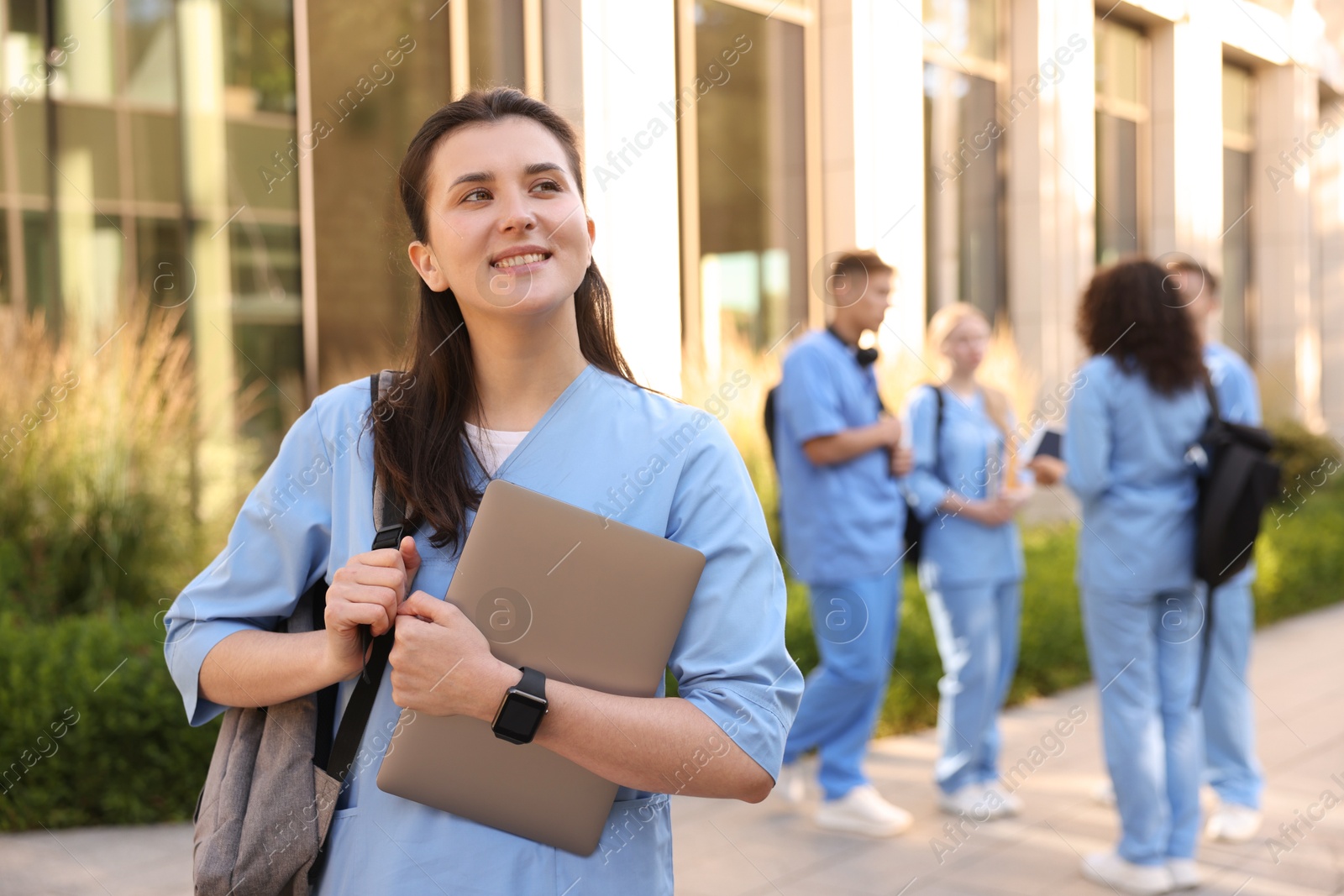 Photo of Happy medical student in uniform with backpack and laptop near building outdoors, selective focus