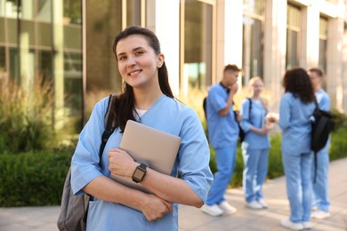 Photo of Happy medical student in uniform with backpack and laptop near building outdoors, selective focus