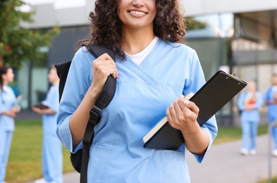 Photo of Medical student in uniform with backpack, clipboard and notebook outdoors, selective focus