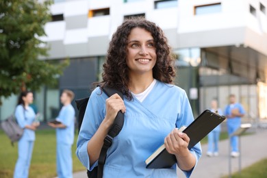Photo of Medical student in uniform with backpack, clipboard and notebook outdoors, selective focus