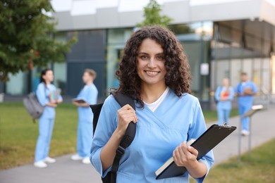 Photo of Medical student in uniform with backpack, clipboard and notebook outdoors, selective focus