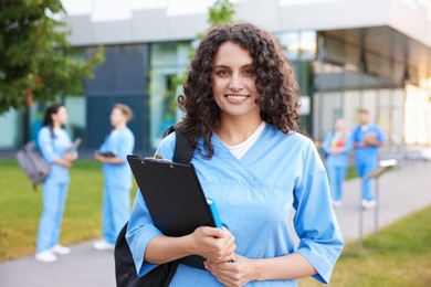 Photo of Medical student in uniform with backpack, clipboard and notebook outdoors, selective focus
